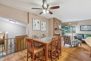 Un coin repas avec une table à plateau en verre, des chaises en bois, un ventilateur de plafond et un mur décoratif en briques.