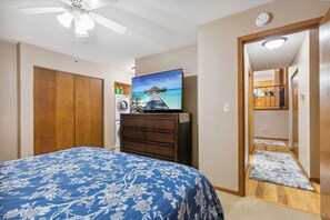 A bedroom with a bed, a ceiling fan, a TV, a wooden wardrobe, and a view of a kitchen through an open door. - Tranquil rural area, near major roads and shops (Lake Elmo)
