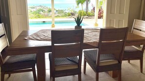 Un espace repas avec vue sur une piscine et l’océan, des chaises en bois et une table.