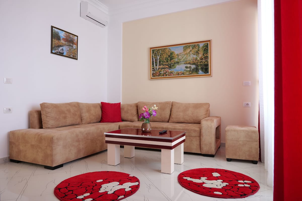 Een woonkamer met een beige bank, een houten salontafel en twee tapijten met een rood patroon.