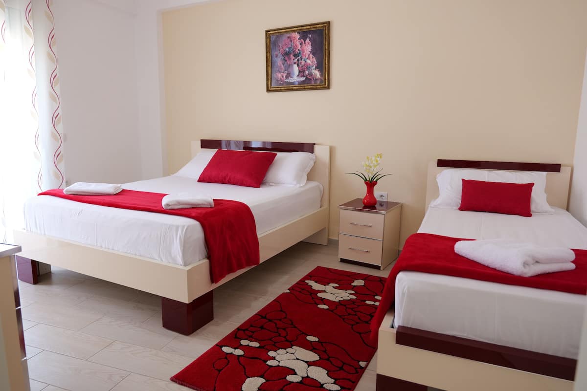Een hotelkamer met twee bedden, elk voorzien van een rode kussens en witte beddengoed. Op de vloer ligt een tapijt met een rood-wit patroon, een klein nachtkastje met een bloemenvaas, en een schilderij aan de muur.