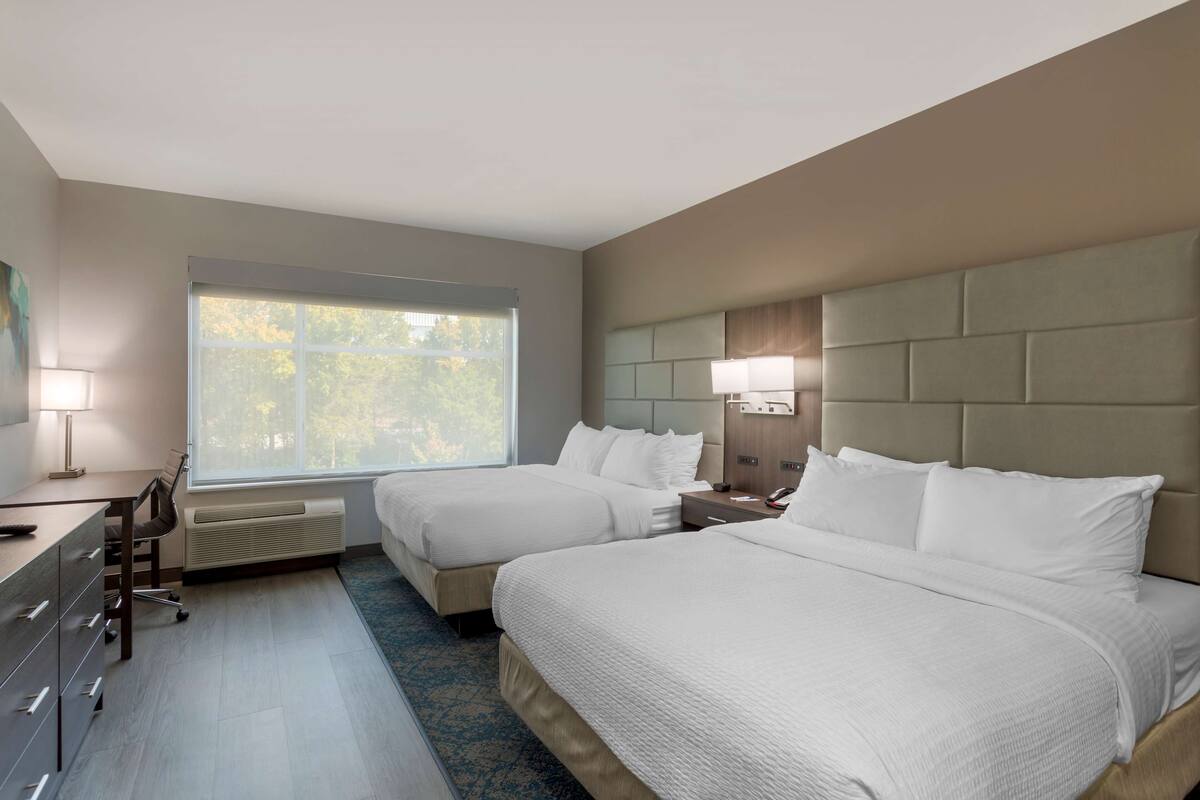A hotel room with two beds, a desk, a chair, a lamp, and a window with a view of trees.