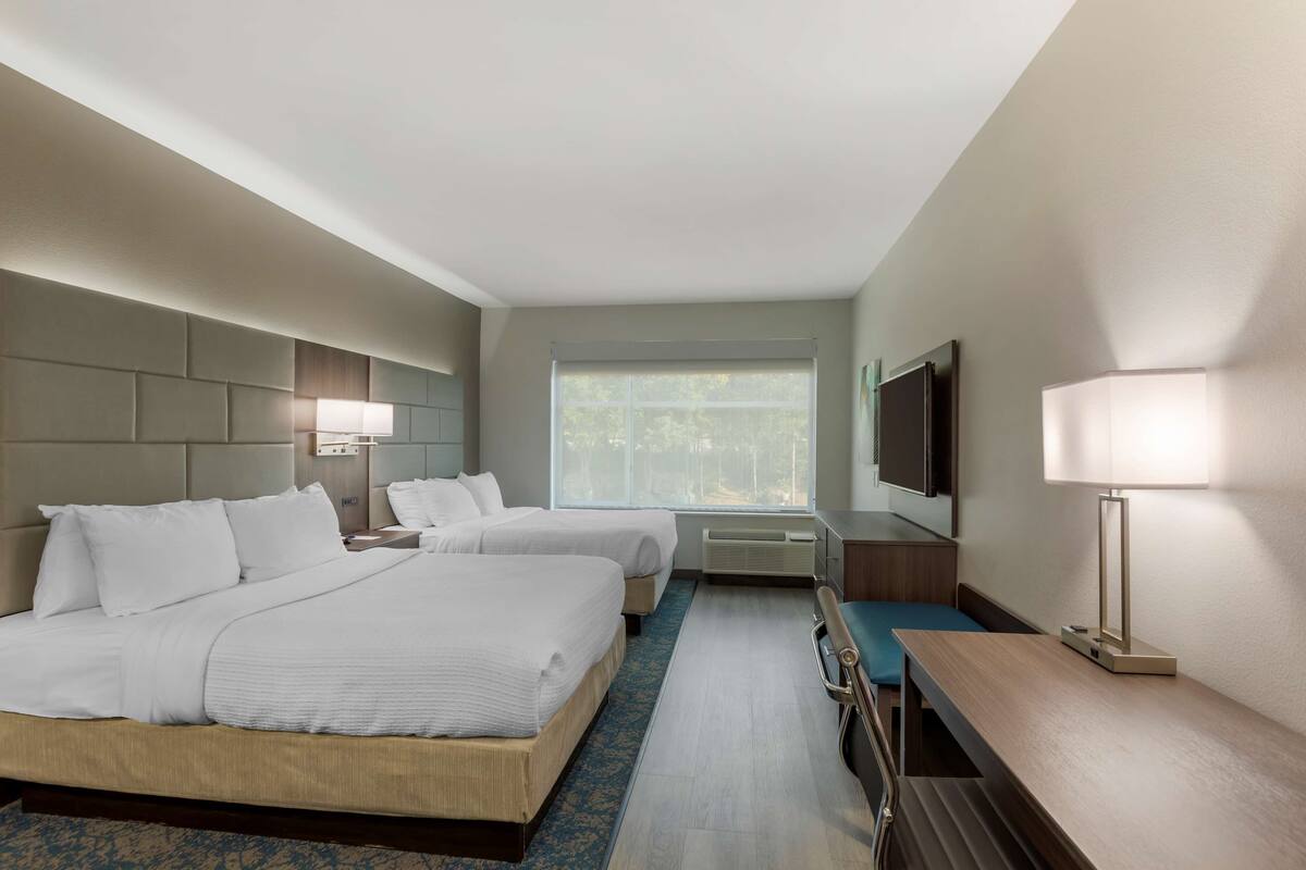 A hotel room with two beds, a desk, a chair, a TV, and a window with a view of trees.