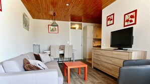 Un salon moderne avec un canapé, une table basse et un téléviseur. On y trouve également un coin repas avec des chaises et une table, ainsi qu’une cuisine équipée de placards et d’un téléviseur.