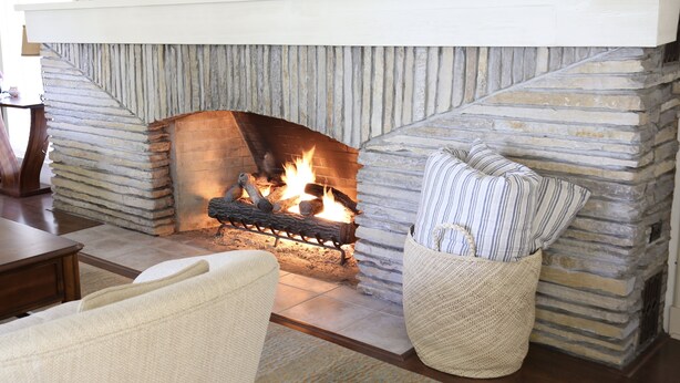 A stone fireplace with a fire, a cushioned chair, and a wooden table in a room with a stone wall. - Mushroom House, Earl Young’s Personal Residence overlooking Lake MI! New AC!  (Charlevoix)