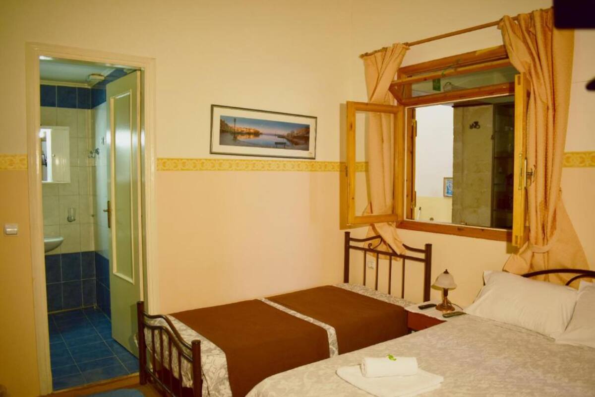 Une chambre avec un lit simple, une table de chevet avec une lampe, une fenêtre avec des rideaux, une porte donnant sur une salle de bain et un tableau encadré au mur.