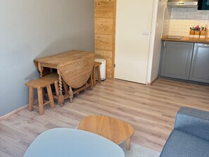 A compact living space with a wooden dining table and benches, a small round table, a grey sofa, and a kitchenette area with cabinets and a countertop.