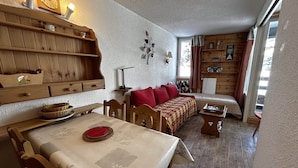 A cozy room with a wooden dining table, a sofa with red cushions, and a bookshelf. - Comfortable studio for 4 with piste view, ski-in/ski-out access (Les Menuires)