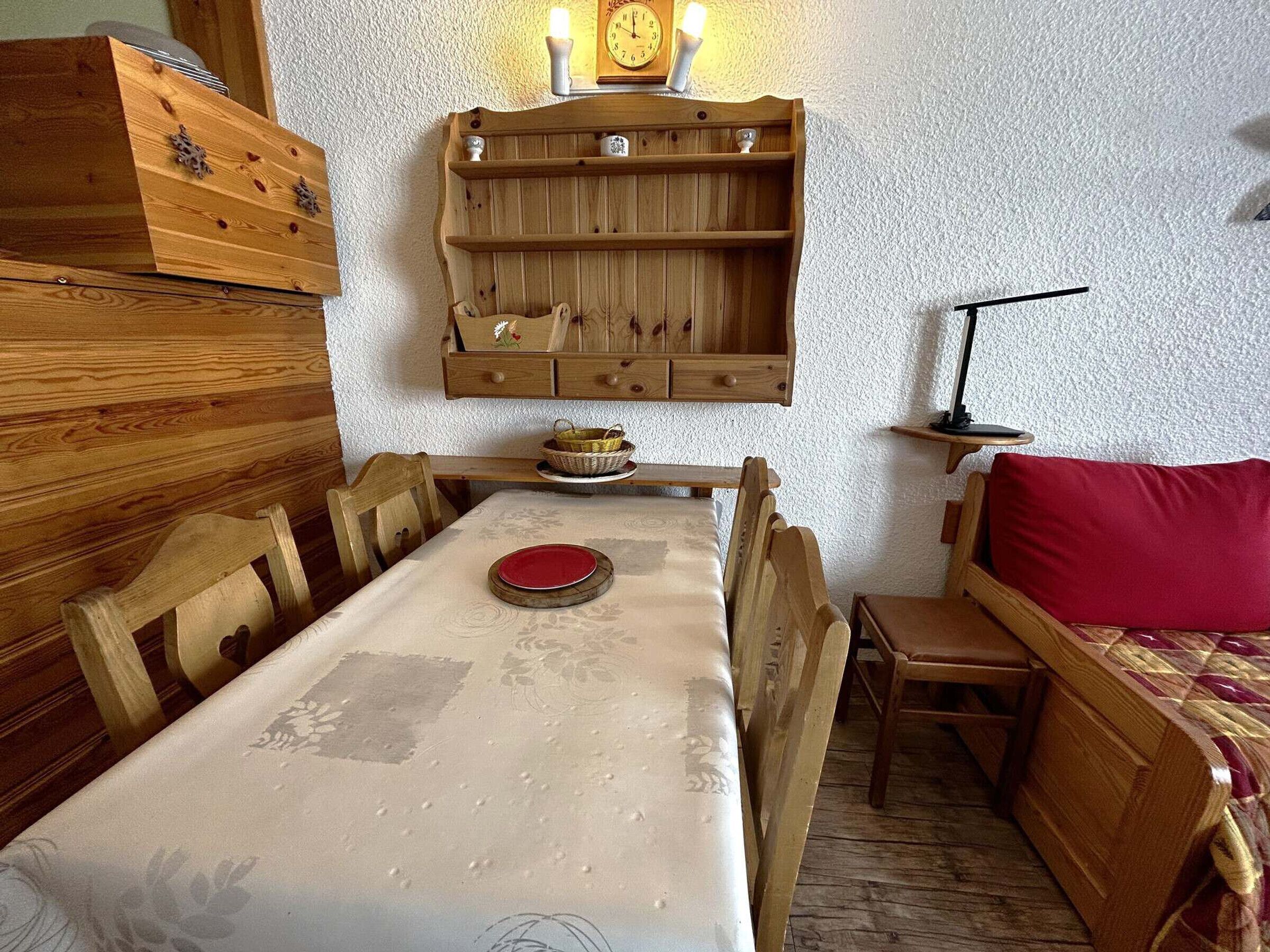 Ett mysigt rum med ett matbord och stolar i trä, en röd soffa, ett träskåp och en vägghylla med en klocka och ett ljus.
