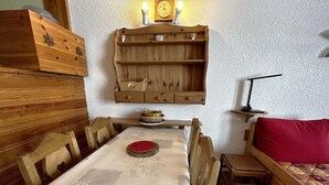 A cozy room with a wooden dining table, chairs, a red sofa, a wooden cabinet, and a wall-mounted shelf with a clock and candle. - Comfortable studio for 4 with piste view, ski-in/ski-out access (Les Menuires)