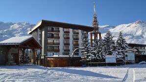 A snow-covered hotel with a prominent tower, surrounded by snow-laden trees and mountains in the background. - Comfortable studio for 4 with piste view, ski-in/ski-out access (Les Menuires)