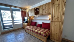 A bedroom with a bed, wooden cabinets, a TV, and a window with curtains. - Comfortable studio for 4 with piste view, ski-in/ski-out access (Les Menuires)