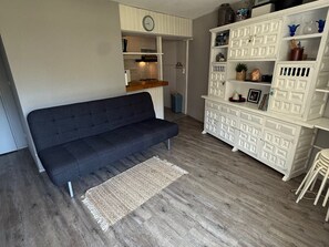 A living room with a dark sofa, a white cabinet with multiple drawers, and a kitchen area visible through an open door.