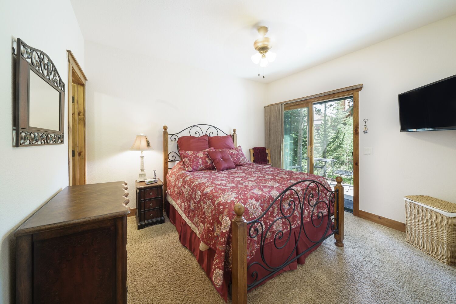 A bedroom with a large bed, a wooden nightstand, a lamp, a dresser, and a television on the wall.