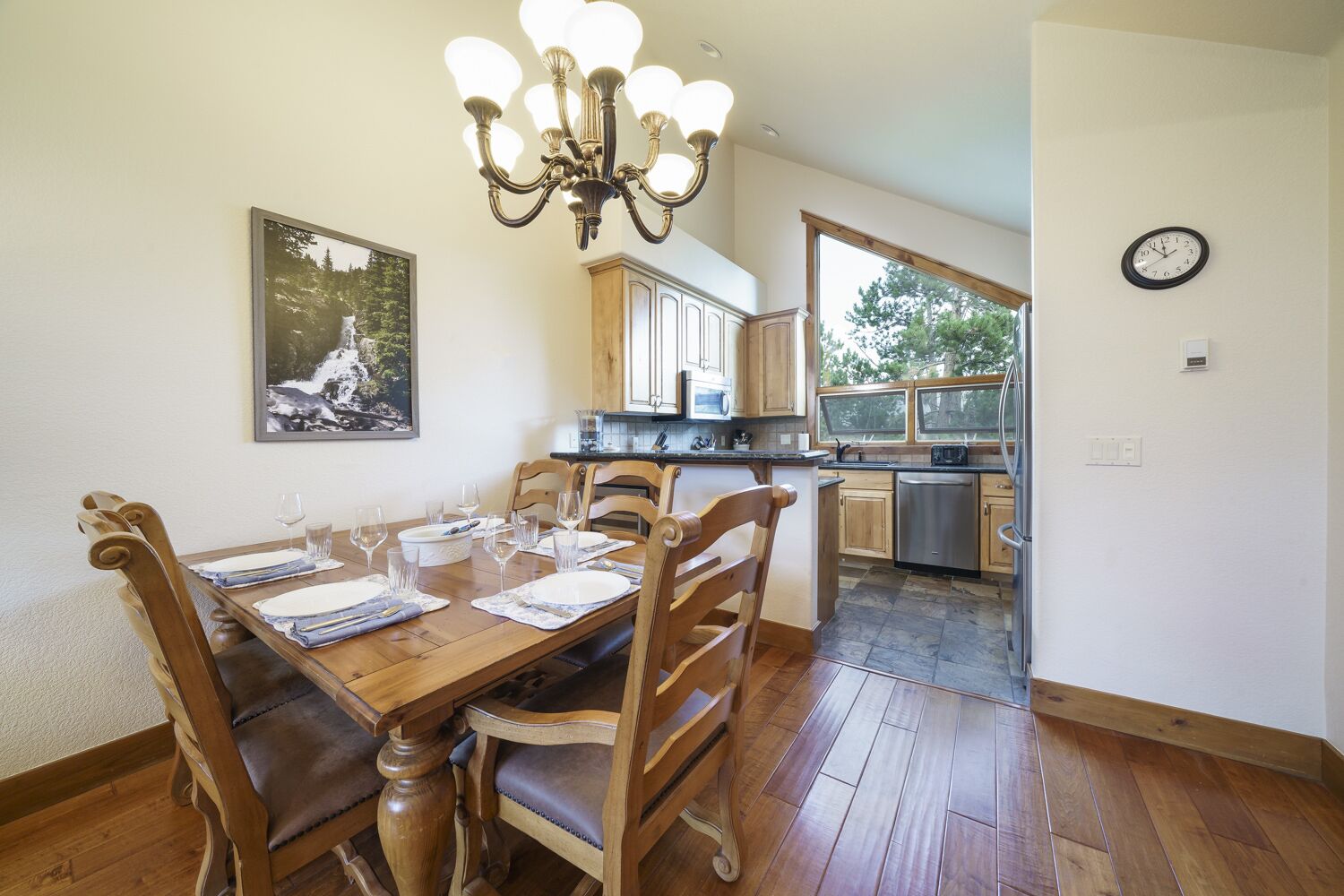 A dining room with a wooden table set for six, a chandelier, and a kitchen in the background.