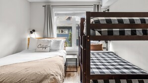 Un dormitorio con literas, una cama individual, una ventana con cortinas y una mesita de noche con lámpara, y vista a una casa desde el exterior.
