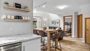 Una cocina moderna con armarios blancos, encimera de mármol y una mesa de comedor de madera con sillas. Hay estantes sobre la encimera con tazas y un letrero de bienvenida.
