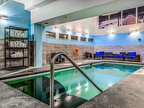 A swimming pool with a glass bottom, a lounge area with blue cushions, and a shelf with folded towels. - Sophisticated Mountain Condo – Pool & Hot Tub (Canmore)