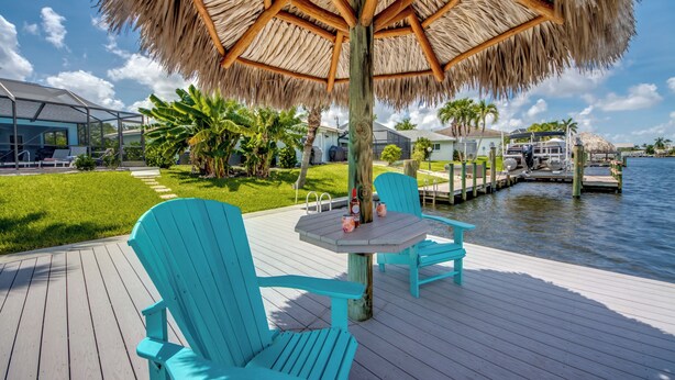 A wooden deck with turquoise chairs and a table, a thatched umbrella, and a view of a dock and water. - Wide Water Views, Hot Tub, Heated pool! 75 reviews.Villa 27 (Cape Coral)