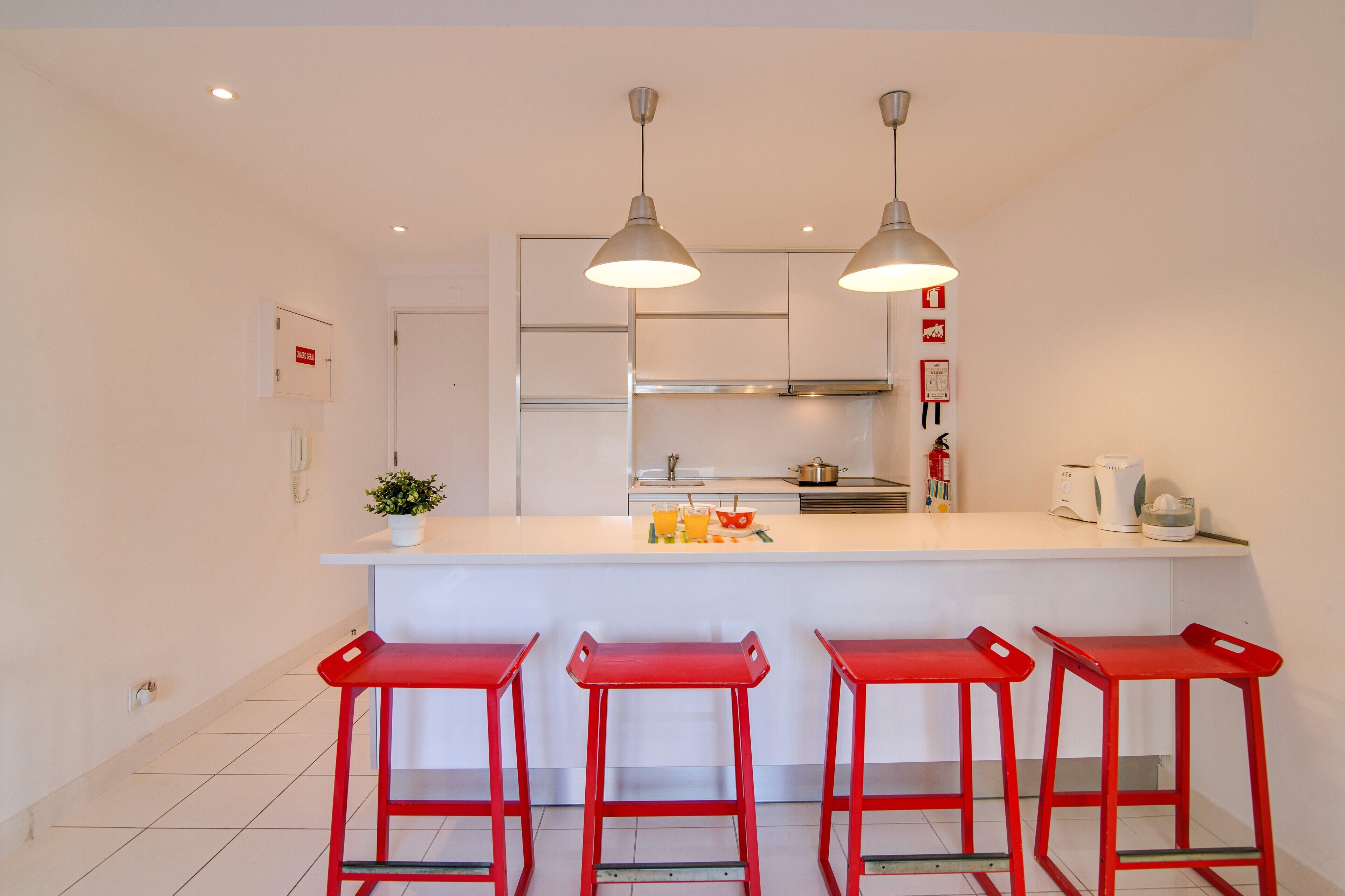 Een moderne keuken met een centraal eiland, rode barkrukken, hanglampen en een wit aanrecht.