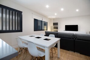 A modern living room with a white dining table, white chairs, a grey sofa, and a flat-screen TV.