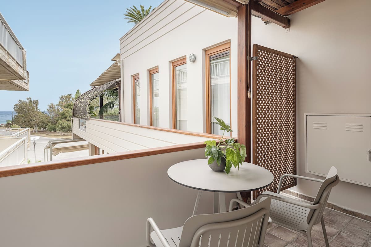 Ein Balkon mit rundem Tisch und zwei Stühlen, einem Topfpflanzen und Blick auf das Meer und die Gebäude.