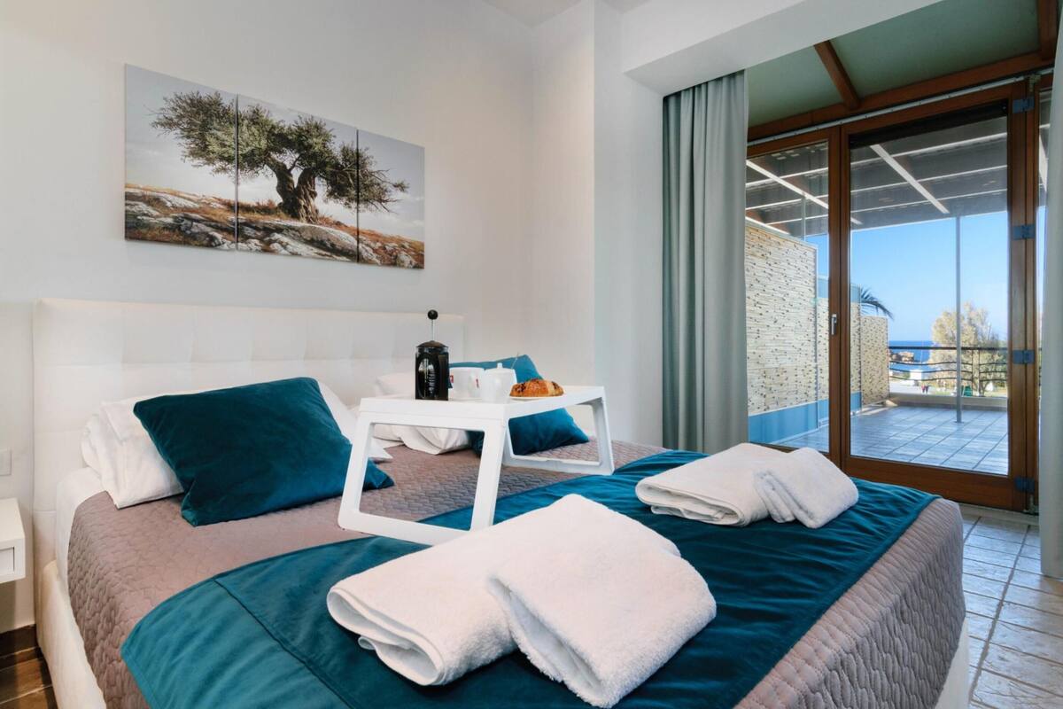 Ein Hotelzimmer mit einem Bett, einem kleinen Tisch mit Tasse und Gebäck, und Blick ins Freie durch ein großes Fenster.
