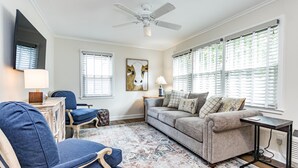 A living room with a sofa, armchair, and a coffee table. There is a ceiling fan, a painting on the wall, and a window with blinds. - Comfortable and Quiet Condo near the Village and NC State (Raleigh)
