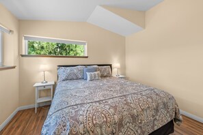 A bedroom with a large bed, two bedside tables, a window with a view of trees, and a sloped ceiling.