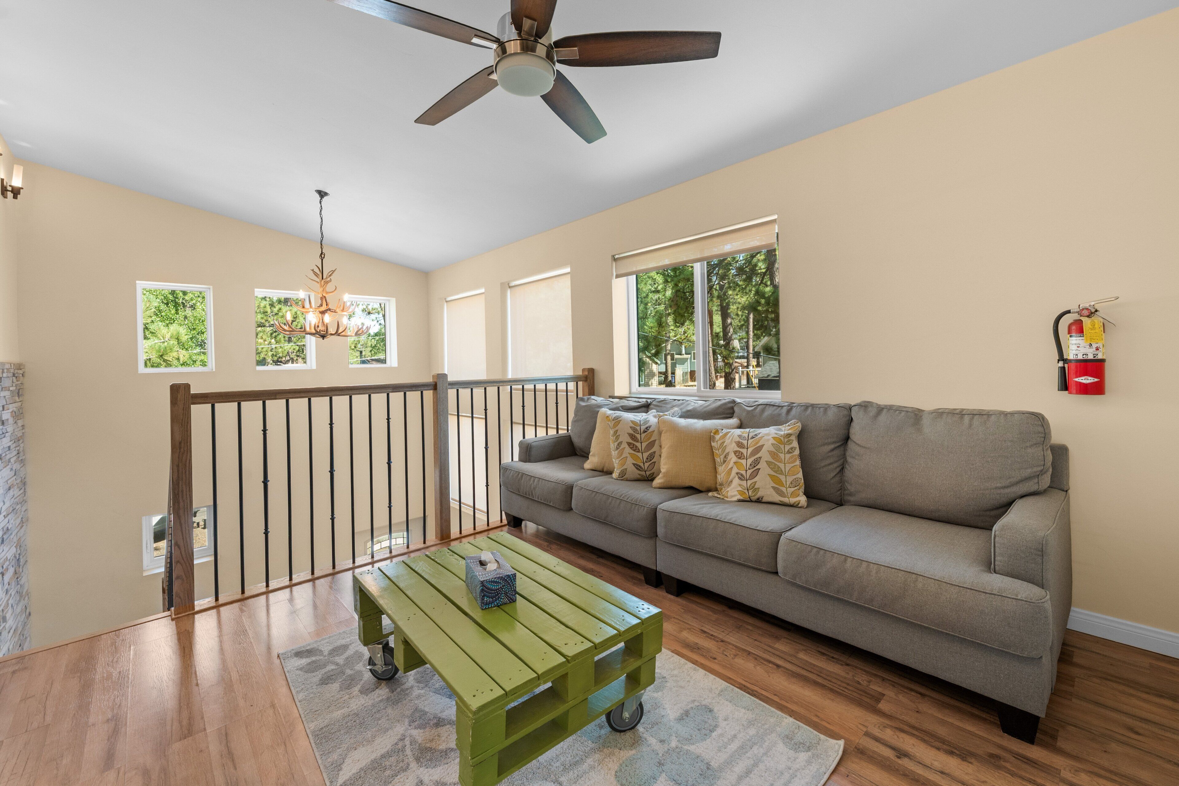A spacious living room with a ceiling fan, a green coffee table, a grey sofa with patterned cushions, and a fire extinguisher mounted on the wall.