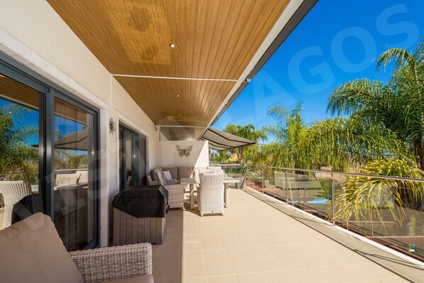 Wraparound terrace with outdoor lounge furniture at Casa Vista Lago, a holiday townhouse in Lagos, Algarve. Features wooden slatted ceiling, glass railings, palm trees, views over the swimming pool and sliding doors to the living area.