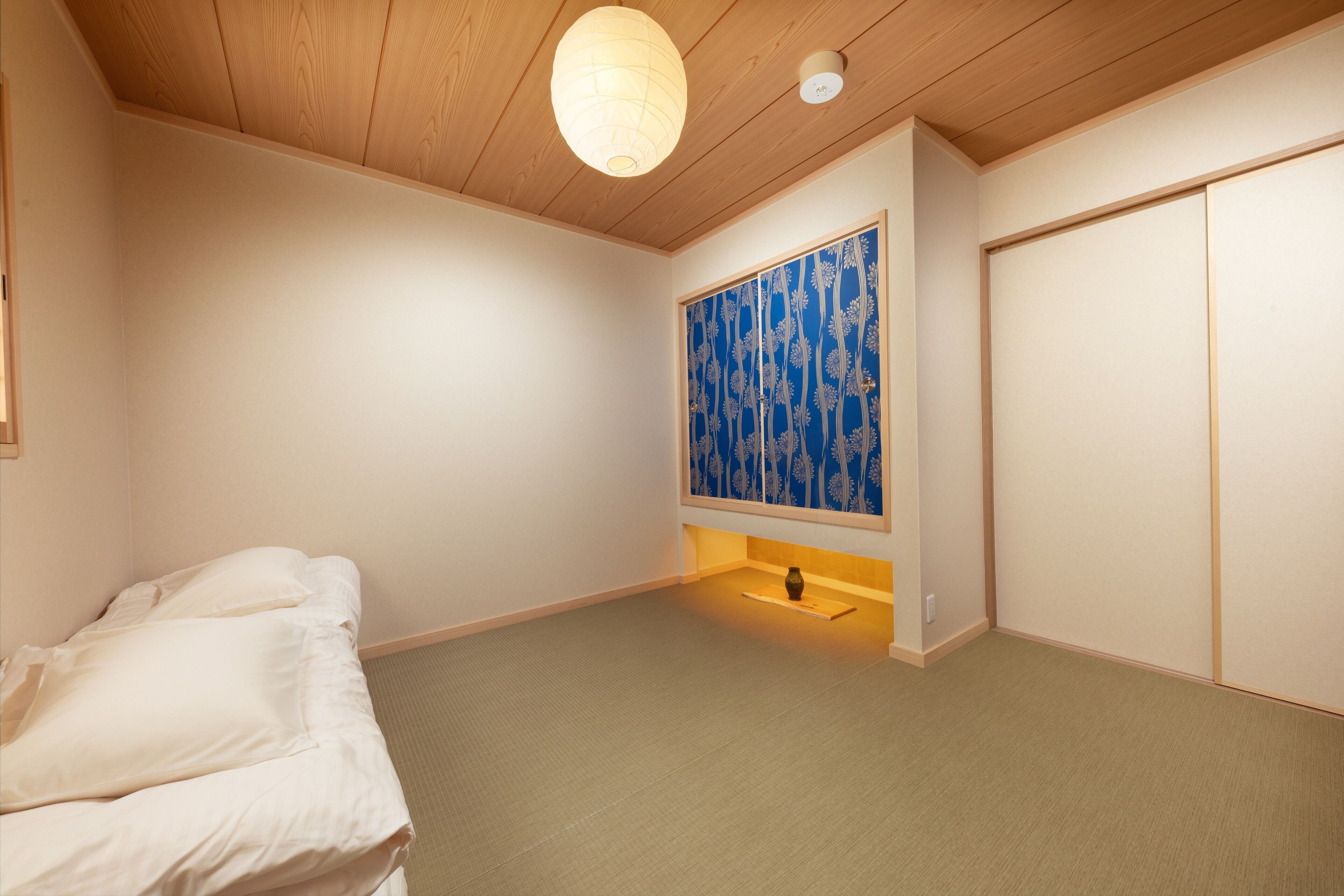A bedroom with a bed, a wall-mounted shelf with a vase, a blue patterned artwork, and a sliding door.