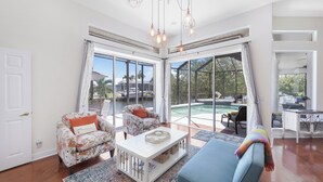 A spacious living room with a large glass door leading to a pool area, a white coffee table, and floral-patterned armchairs. - Waterfront Home on Key Lot | Pool & Sundeck (Marco Island)