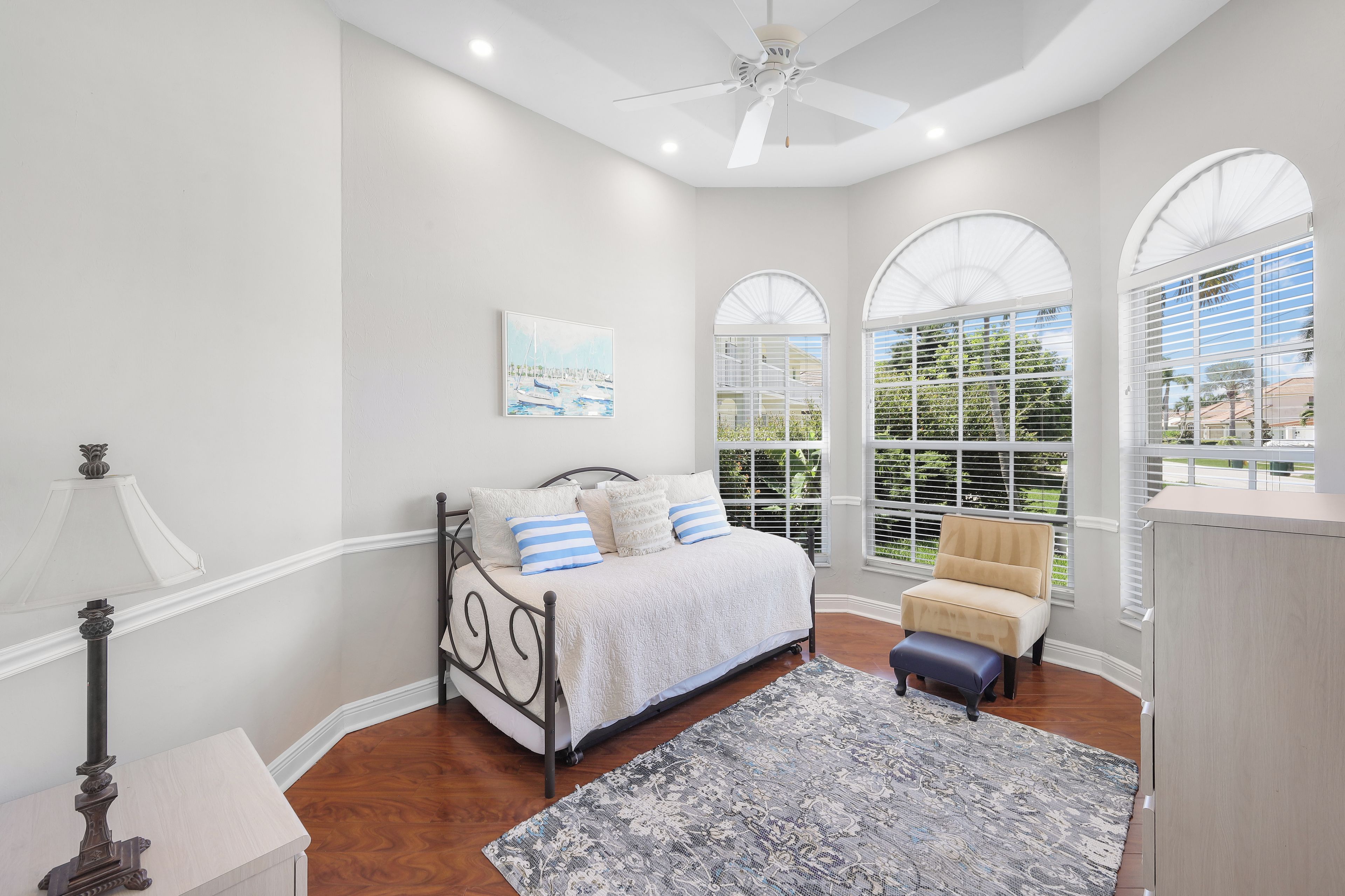 A bedroom with a bed, a nightstand, a lamp, a ceiling fan, and large arched windows.