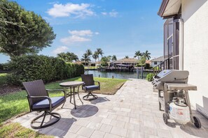 An outdoor patio with a grill, seating, and a view of a water body and buildings in the background. - Waterfront Home on Key Lot | Pool & Sundeck (Marco Island)