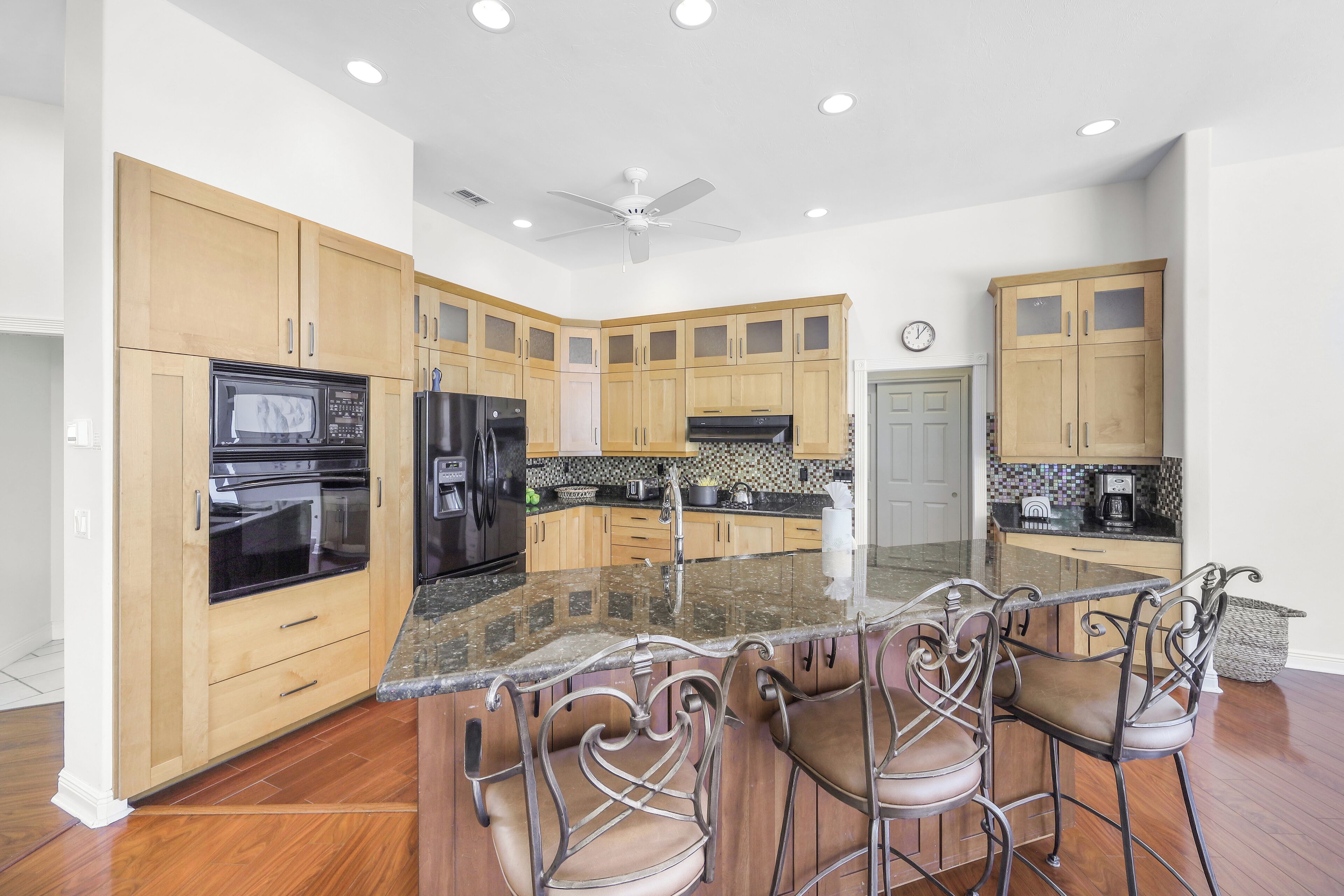 A modern kitchen with wooden cabinets, a black refrigerator, a microwave, and a stove. There is a bar area with metal chairs and a granite countertop.