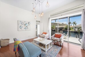 Ein geräumiges Wohnzimmer mit großem Fenster, einem farbenfrohen abstrakten Gemälde sowie Blick auf einen Pool und Palmen.