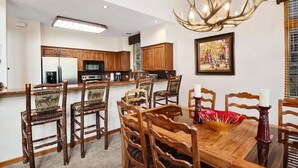 A dining area with a wooden table and chairs, a chandelier, and a kitchen in the background. - Enclave #217 5 Bedroom Home by RedAwning (Avon)