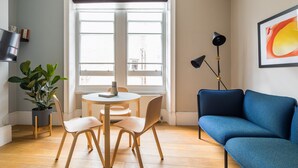 A modern living room with a blue sofa, a round table with chairs, a potted plant, and a large window with blinds. - Vibrant Covent Garden 1BR - Netflix & Nespresso (London)