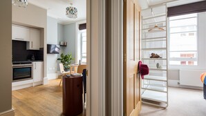 A modern kitchen with white cabinets, a black countertop, and a built-in oven. There is a dining area with a table and chairs, a potted plant, and a TV mounted on the wall. A white ladder with a red hat hangs on the door. - Vibrant Covent Garden 1BR - Netflix & Nespresso (London)