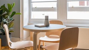 A round table with a book and a vase, surrounded by two chairs, in a room with a large window and a potted plant. - Vibrant Covent Garden 1BR - Netflix & Nespresso (London)