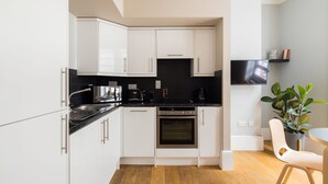 A modern kitchen with white cabinets, a black countertop, and stainless steel appliances. There is a dining area with a table and chairs, and a potted plant. - Vibrant Covent Garden 1BR - Netflix & Nespresso (London)
