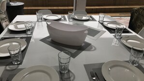 A dining area with a table set for four, including plates, glasses, and cutlery. A large white bowl is placed in the center. The room has a view of a swimming pool and lounge chairs outside. - House on the south coast with optional heated pool. (Mongaguá)