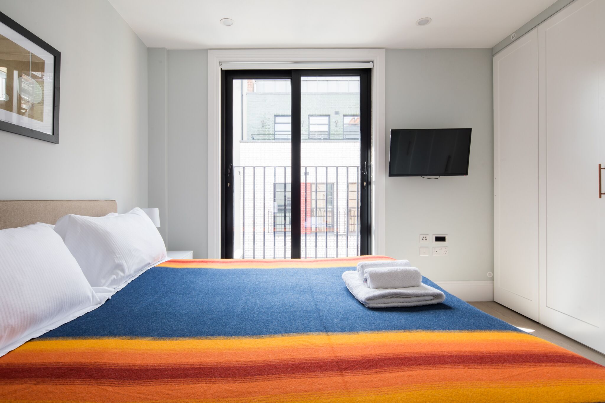A hotel room with a bed, a TV mounted on the wall, a balcony door, and a view of buildings outside.