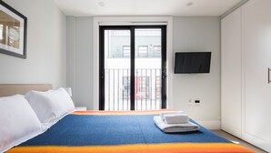 Une chambre d’hôtel avec un lit, un téléviseur fixé au mur, une porte-fenêtre donnant sur un balcon et une vue sur les bâtiments environnants.