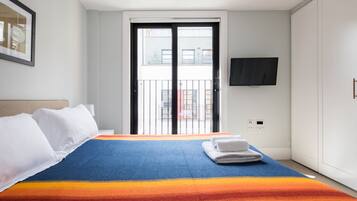 A hotel room with a bed, a TV mounted on the wall, a balcony door, and a view of buildings outside.