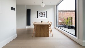 Un comedor moderno con una mesa y sillas de madera, una lámpara colgante, y vistas a una pared de ladrillo y plantas afuera.