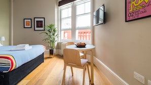 Ein Hotelzimmer mit einem Bett, einem kleinen Tisch mit einer Schale, einem Sessel, einer Zimmerpflanze, einem an der Wand befestigten Fernseher und gerahmten Bildern an der Wand.