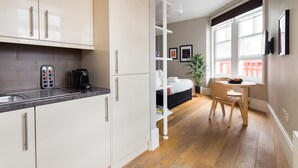 Eine moderne Küche mit weißen Schränken, einer Kaffeemaschine und einem Spülbecken. Ein Essbereich mit Tisch und Stühlen sowie ein Bett mit weißem Kopfteil sind ebenfalls vorhanden.