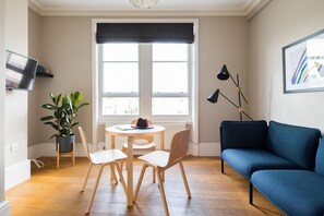 A modern living room with a blue sofa, wooden chairs, a small table, and a potted plant. - Lovely Covent Garden 1BR - Netflix & Nespresso (London)